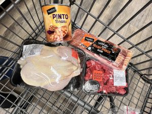 Beans, a whole chicken, a pack of bacon, and beef for stew in a cart. The 6-to-1 grocery shopping method allows for 4 proteins.
